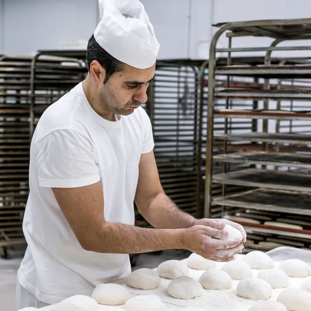 Baker in whites and a hat shaping dough beside bakery racks
