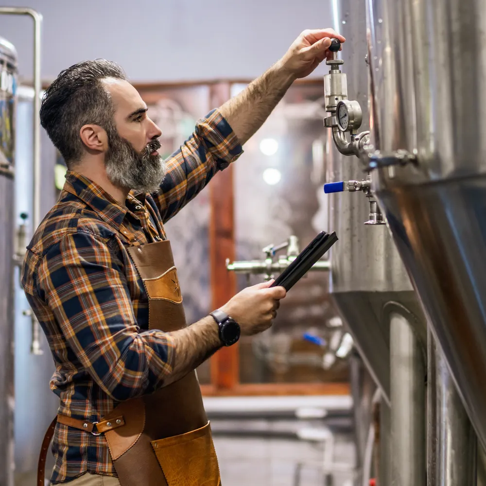 Brewer in a work apron checking a gauge on a stainless steel fermentation tank