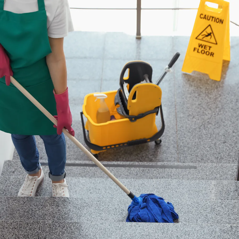 Cleaner in an apron and rubber gloves mopping stone steps beside a wet floor sign