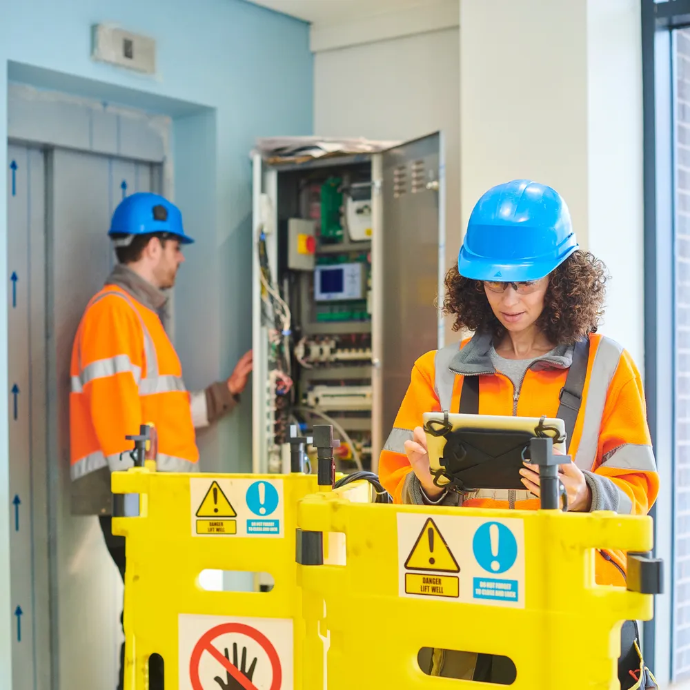 Lift engineers in hi-vis working on a lift control panel behind safety barriers