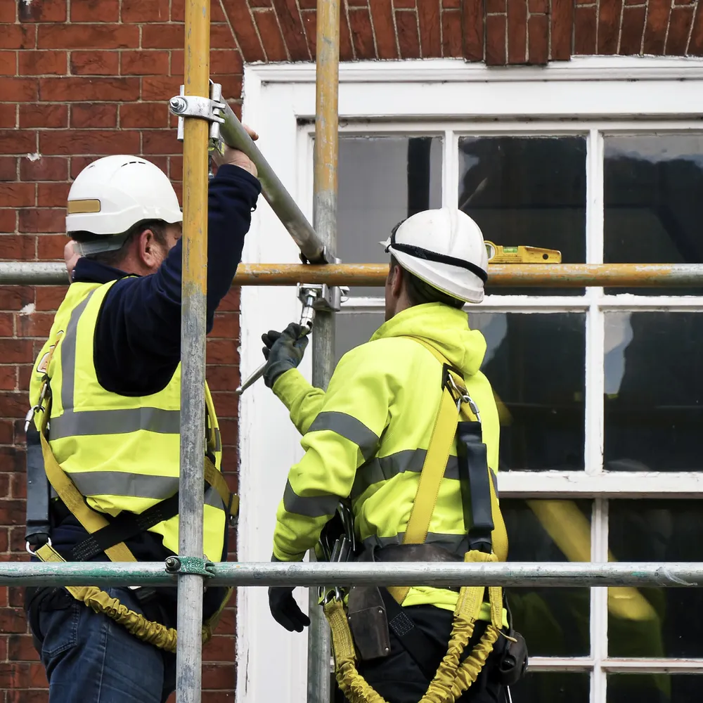Two scaffolders in harnesses and hi-vis fixing scaffold tube on a brick elevation