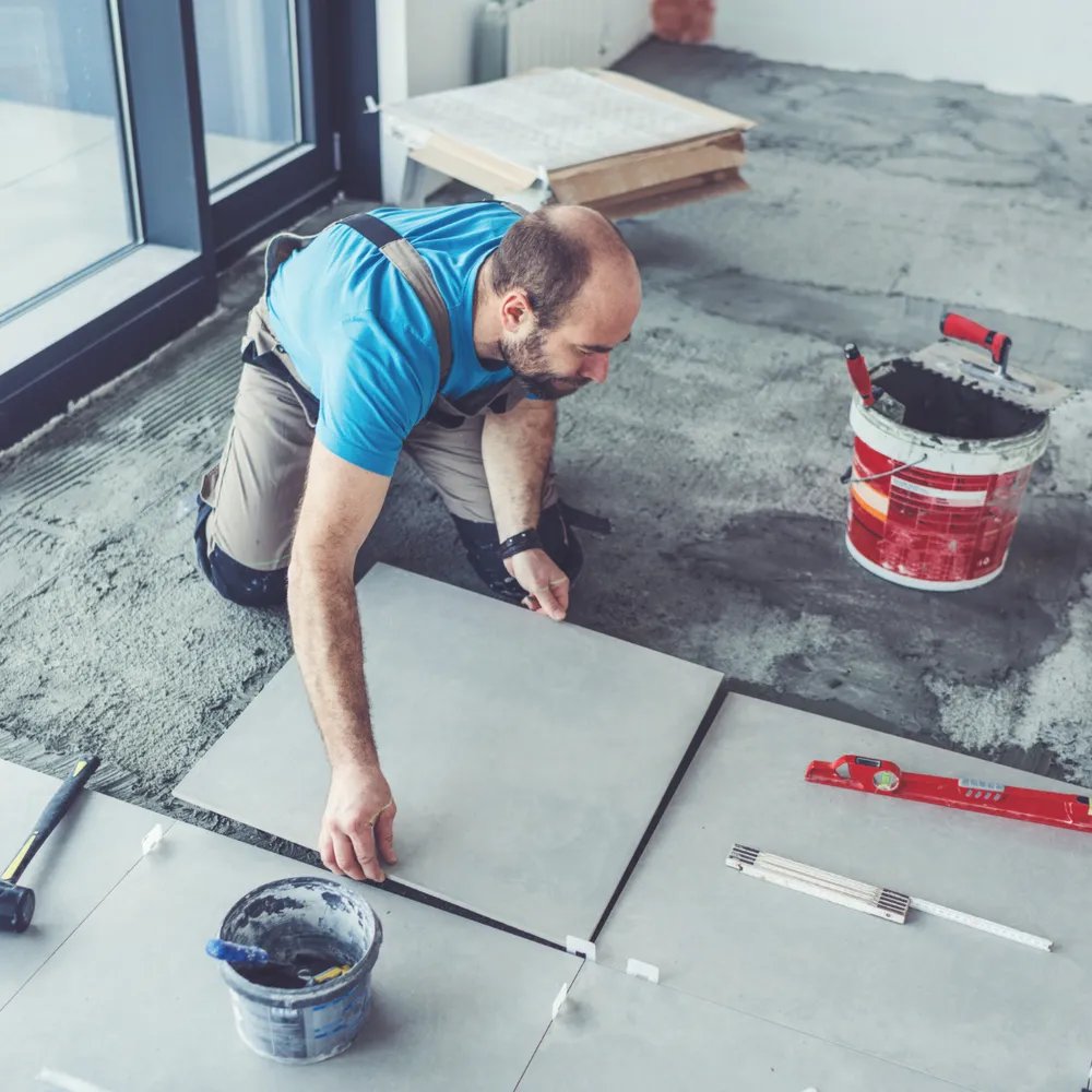 Tiler in bib-and-brace kneeling to lay large floor tiles